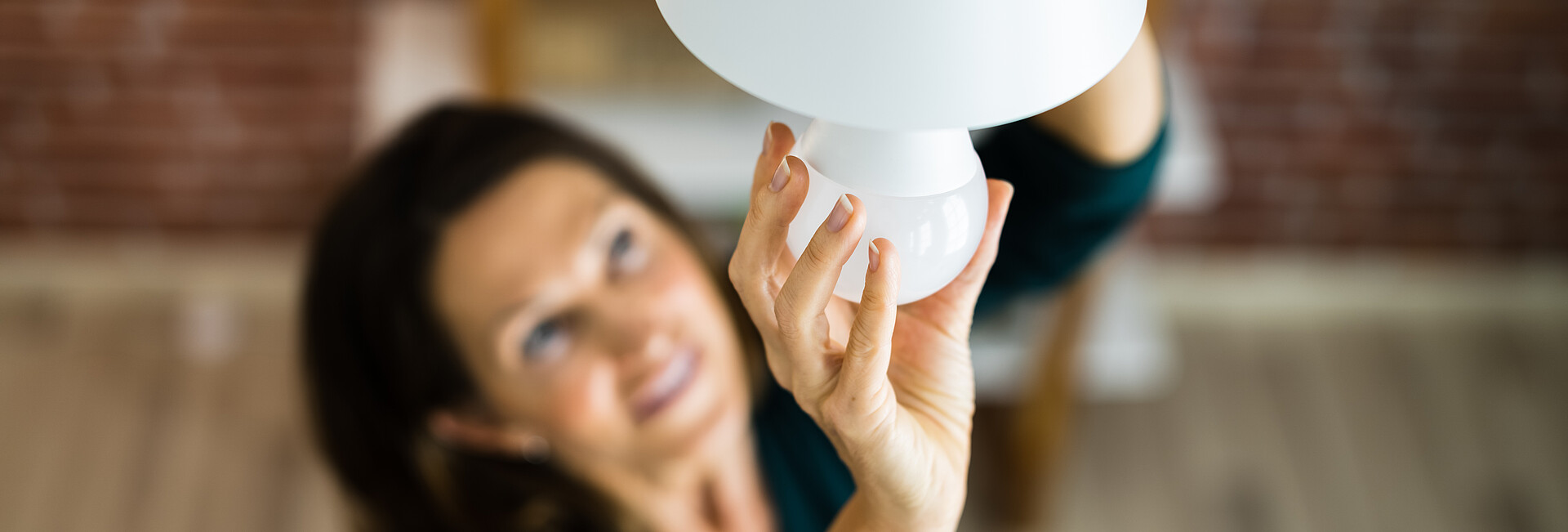 Man blickt von oben auf eine schwarzhaarige mittelalte Frau, die eine neue LED-Birne in die Lampe eindreht: Stichpunkt:LED-Lampe