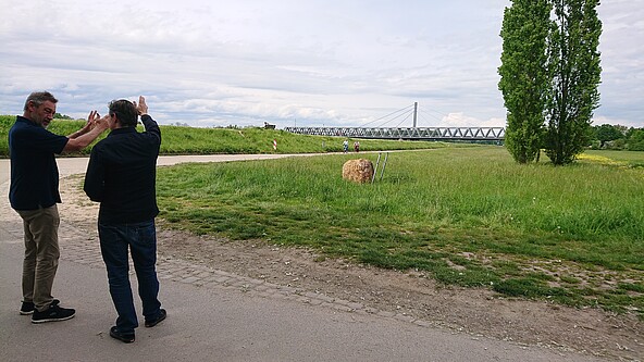 Zwei Männer stehen am Damm in der Nähe des Hofguts Maxau und zeigen im Gespräch mit den Händen entlang des Dammes. Rechts ist landwirtschaftliche Nutzfläche und zwei Pappeln. Hinten sieht man zwei Radfahrer und die Rheinbrücke. 