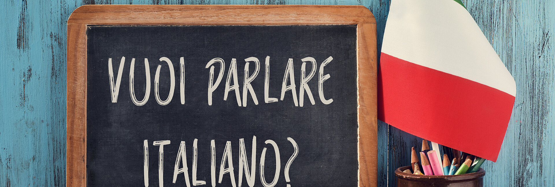 Kreidetafel mit Beschriftung "vuoi parlare italiano?" vor türkisfarbener Wand. Davor ein Gefäß mit Stiften und spanischer Fahne. Rechts neben dem Gefäß liegen zwei übereinander gestapelte Bücher. 