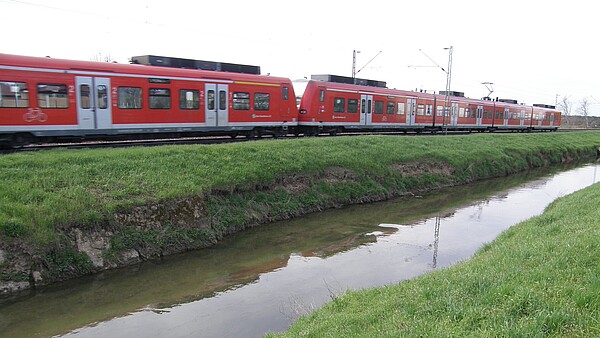 Auf dem Bild sieht man im Vordergrund den monotonen Gewässerlauf des Leimbachs und im Hintergrund die unmittelbar angrenzende parallel verlaufende Bahntrasse auf der gerade ein Zug fährt.