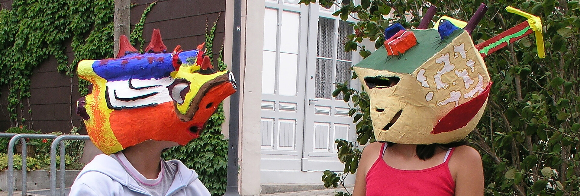 Schülerin und Schüler im Kunstunterricht, die sich gegenübersitzend ansehen und dabei selbst gestaltete bunte Masken tragen
