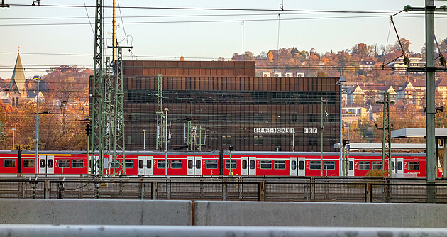 Bild zeigt eine rote S-Bahn mit Gleisen und Oberleitungen im Vordergrund und einem großen, rotem Gebäude im Hintergrund