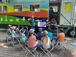 Eine Gruppe von Kindern sitzt im Halbkreis neben dem Ökomobil und schaut auf einen Bildschirm, neben dem sich die Ökomobilistinnen befinden. Gezeigt wird eine lila Blüte in Nahaufnahme.