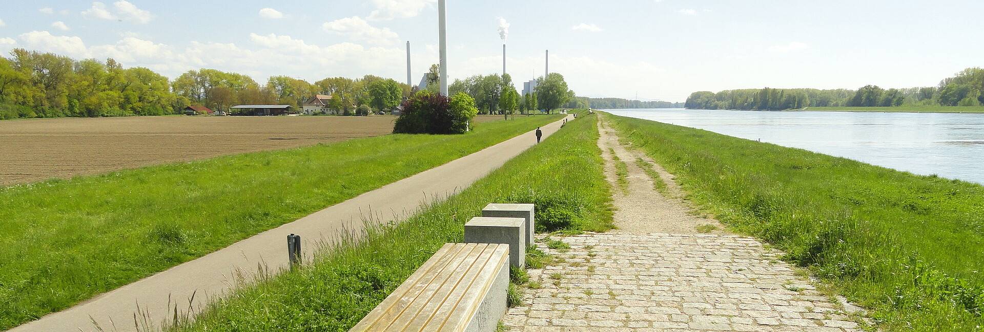 Auf dem Bild sieht man rechts den Rhein und links Wiesen und das Hofgut Maxau. Entlang des Rheins sieht man den unbefestigten Weg auf dem Rheindamm (rechts) sowie den befestigten Rad- und Fußweg am Dammfuß (links), mit Fußgänger. In Hintergrund befindet sich das Rheinhafen-Dampfkraftwerk, im Vordergrund einige Sitzgelegenheiten. Mittig, allerdings oben angeschnitten, befindet sich ein Windrad. 