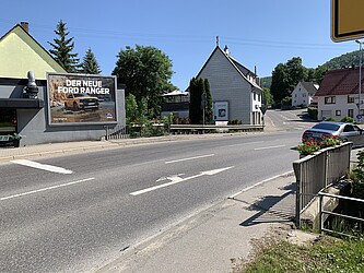 Eine Straße in einem Ort, am rechten Bildrand ist ein Brückengeländer zu erkennen