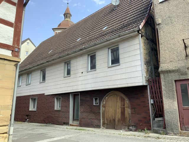 Das Bild zeigt die Frontansicht eines älteren Hauses. Neben der Eingangstür befindet sich noch ein oben abgerundetes Holztor.