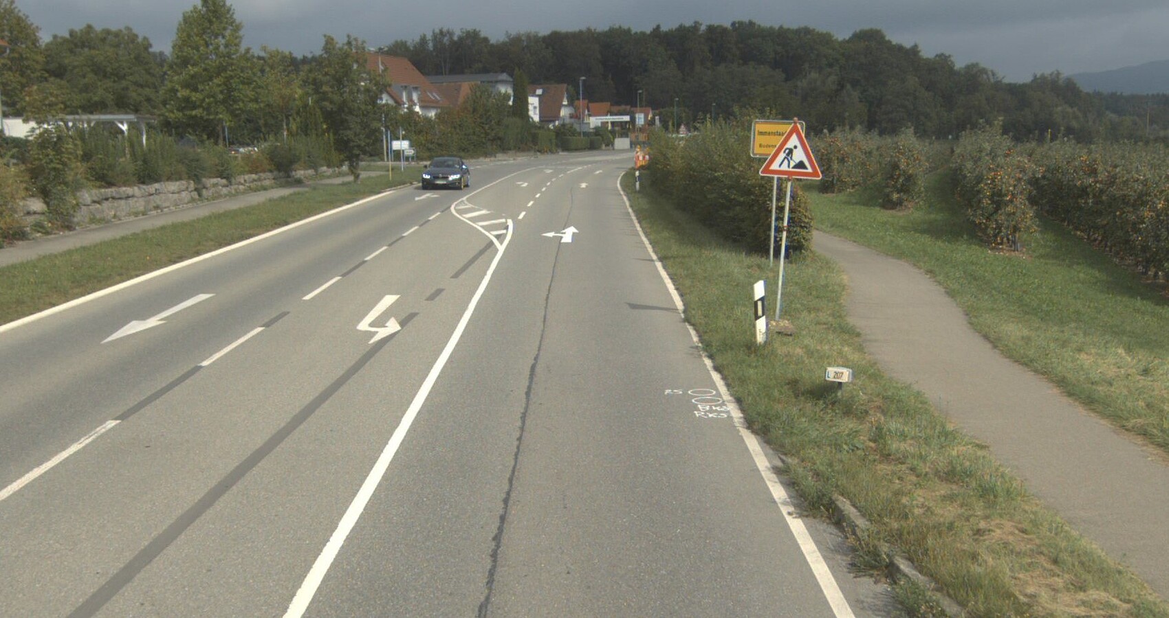 Das Foto zeigt eine Landesstraße mit einer Abbiegespur. Ein Baustellenschild und ein Ortsschild. Links und rechts neben der Straße ein Radweg. Rechts Baumwiesen. Links ein Wohngebiet