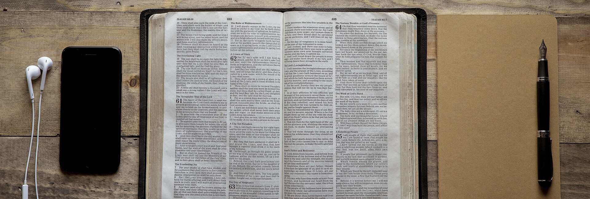 Geöffnete Bibel; rechts daneben ein Notizbuch, auf dem ein Füller liegt. Links von der Bibel liegt ein ein Smartphone mit einem Kopfhörer.