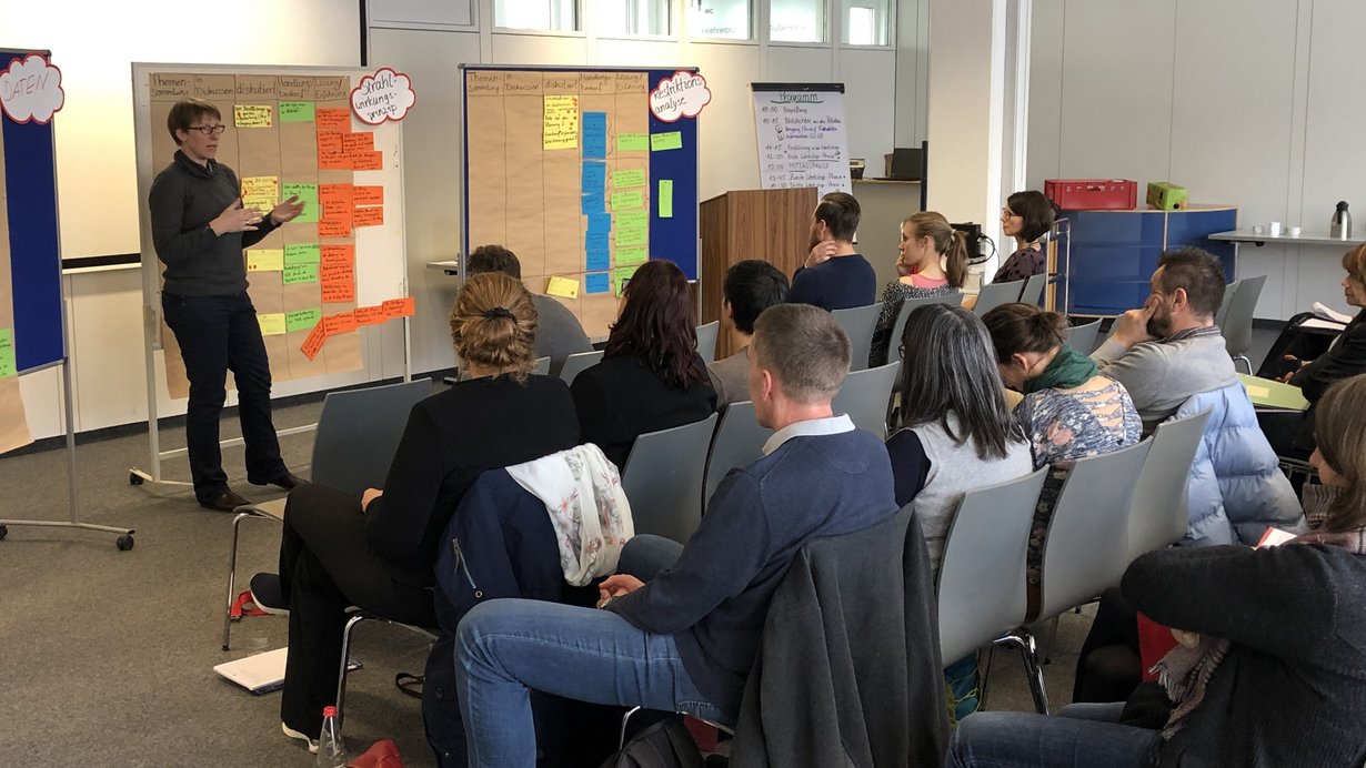 Eine Referentin spricht vor dem Visualiserungsboard und stellt einer Gruppe von Teilnehmenden Ergebnisse einer Gruppendiskussion vor.