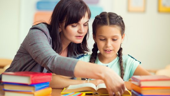 Junge Lehrerin mit Schülerin an einem Schreibtisch mit vielen Büchern darauf