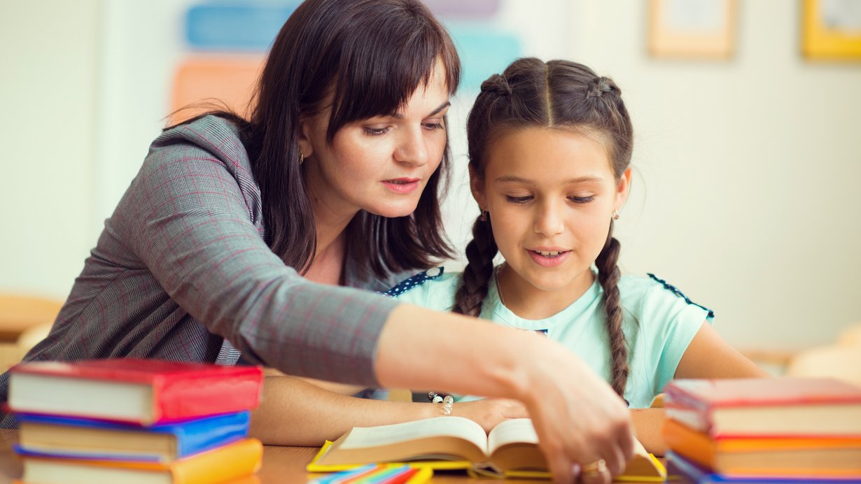 Junge Lehrerin mit Schülerin an einem Schreibtisch mit vielen Büchern darauf