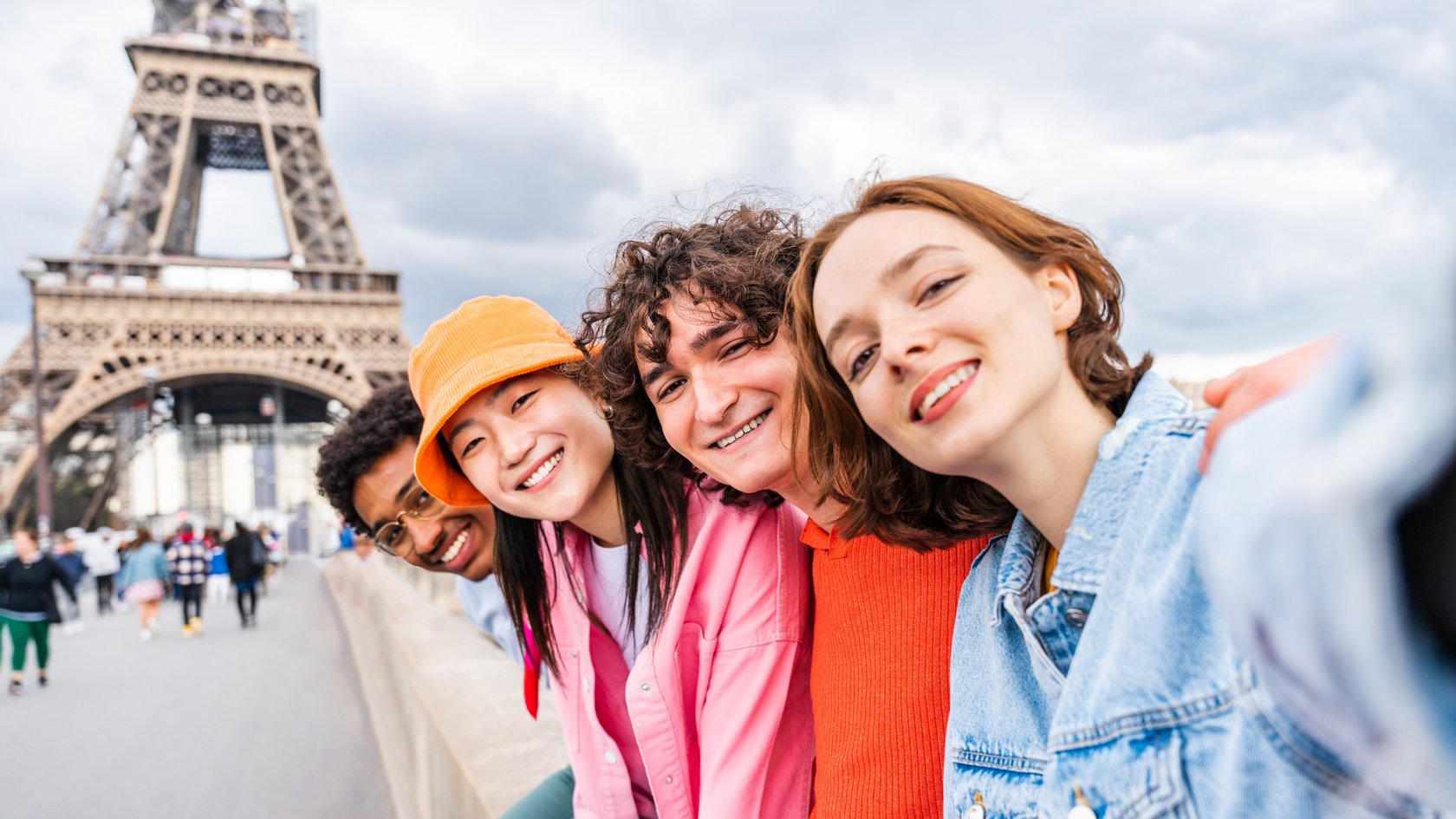 zwei Mädchen und zwei Jungs sitzen in Reihe auf einem Geländer und lächeln in die Kamera. Im Hintergrund sieht man den Pariser Eiffelturm.