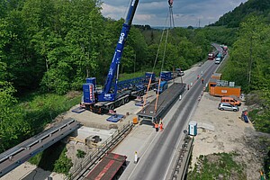 Ein Kran steht neben einer Straße. Er Hebt ein Brückenteil, das direkt neben der Straße liegt, auf circa einen Meter Höhe. Vor dem Kran befinden sich Brücken-Widerlager, auf denen bereits ein Brückenteil abgelegt ist. 