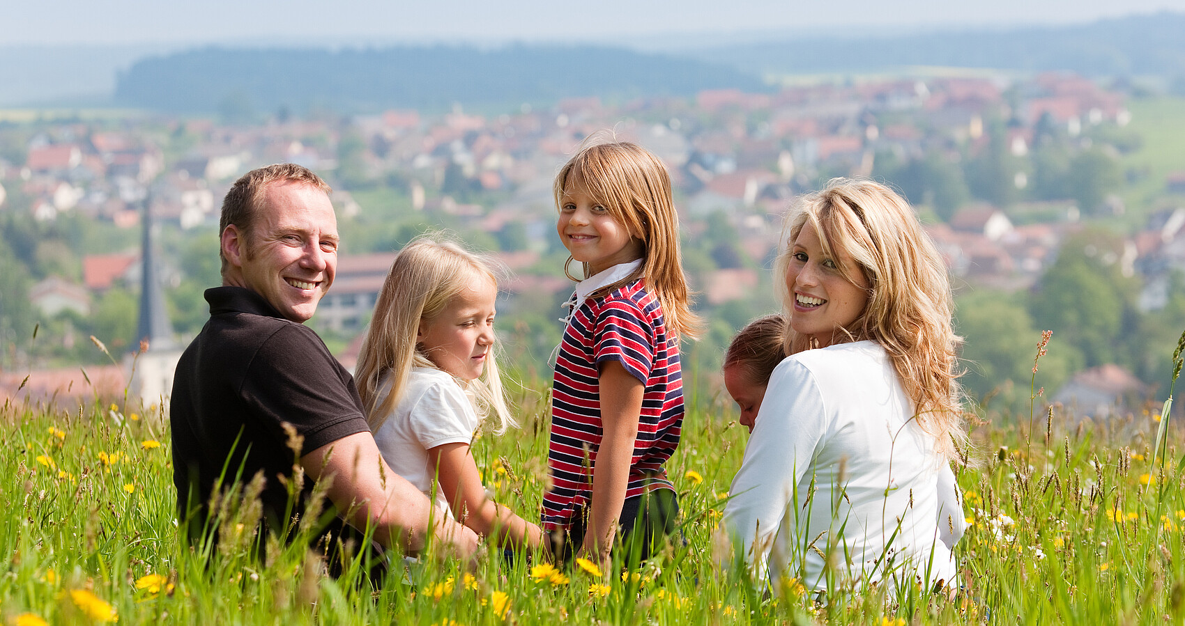 Man sieht eine Familie mit zwei Kindern auf einer Wiese im Frühling oder Sommer mit Blick auf ein Dorf
