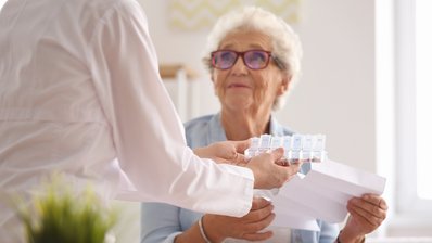 Eine ältere Dame sitzt im Sessel und bekommt von einer Helferin Medikamente zur Einnahme überreicht.