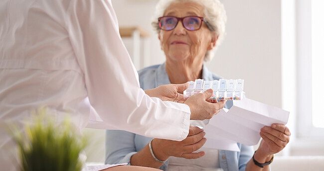 Eine ältere Dame sitzt im Sessel und bekommt von einer Helferin Medikamente zur Einnahme überreicht.