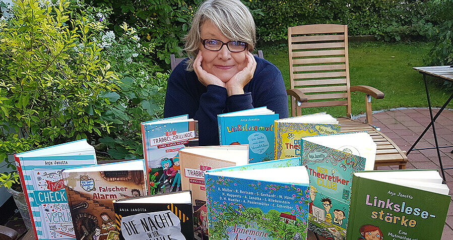 Das Bild zeigt eine Frau auf einer Terrasse. Sie sitzt an einem Tisch und hat die ihr Kinn in die Hände gestützt. Links von ihr ist ein Busch, auf dem Tisch vor ihr stehen mehrere Bücher. 