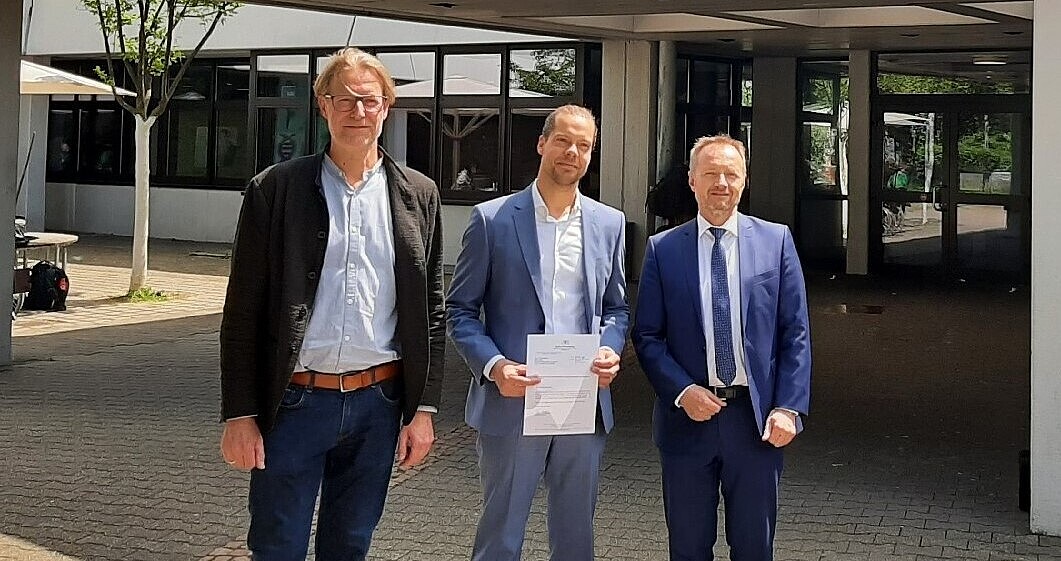 Bild zeigt Dr. Thomas Hölz und Armin Dunz bei der Überreichung des Bestellungsschreibens an Daniel Meier vor dem Gustav-Stresemann-Gymnasium