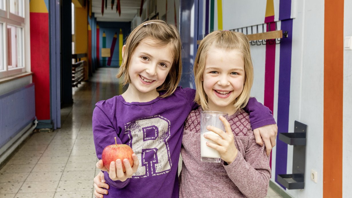 Zwei lachende Mädchen halten einen Apfel und Milch in der Hand
