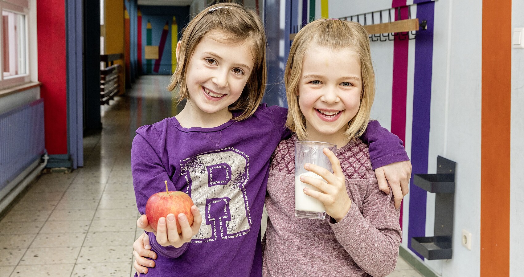 Zwei lachende Mädchen halten einen Apfel und Milch in der Hand