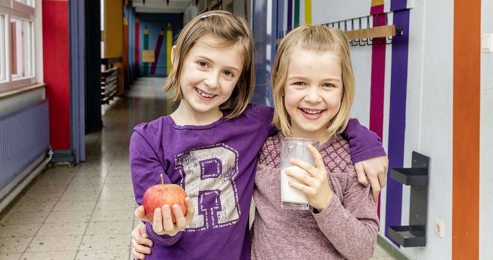 Zwei lachende Mädchen halten einen Apfel und Milch in der Hand