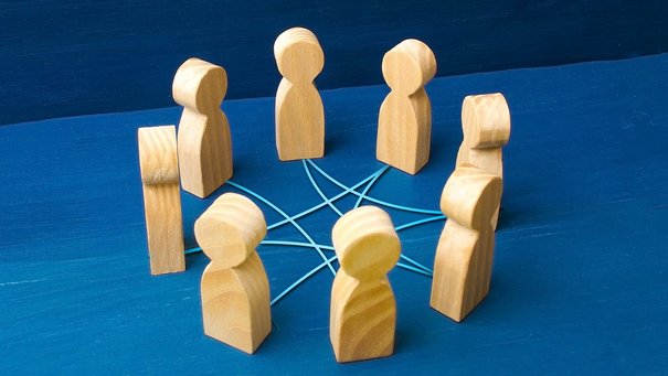 Acht Holzmännchen stehen auf einer blauen Fläche im Kreis und sind über Fäden verbunden.