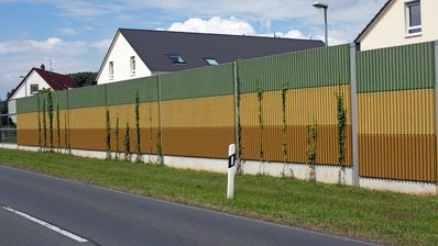 Lärmschutzwände entlang einer Wohnsiedlung, Im Vordergrund sieht man eine nicht befahrene Straße, hinter der Lärmschutzwand sind die Giebel der Häuser zu sehen
