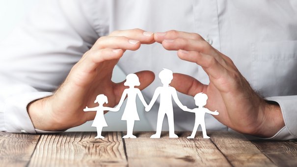 Eine Familie aus Papier, darüber formt ein Mensch ein Dach mit seinen Händen