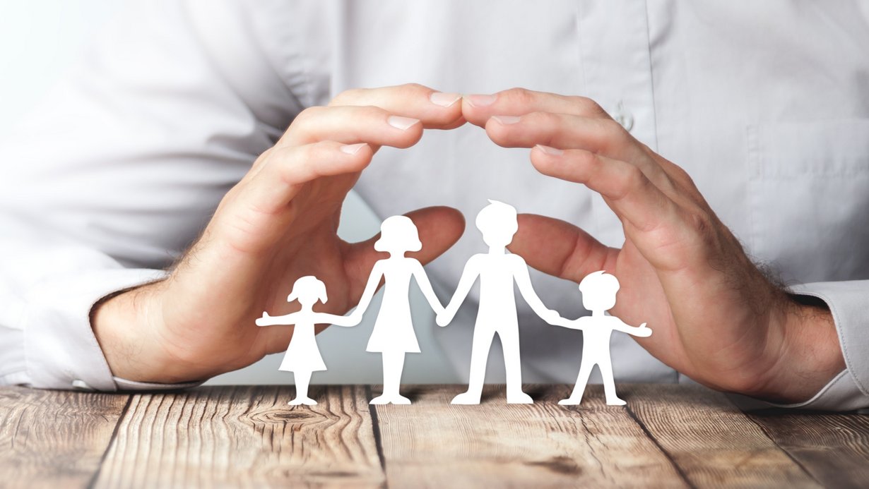 Eine Familie aus Papier, darüber formt ein Mensch ein Dach mit seinen Händen