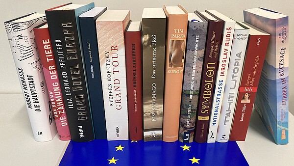 14 Bücher stehen aufrecht mit dem Rücken zum Betrachter, davor liegt eine Europa-Fahne.