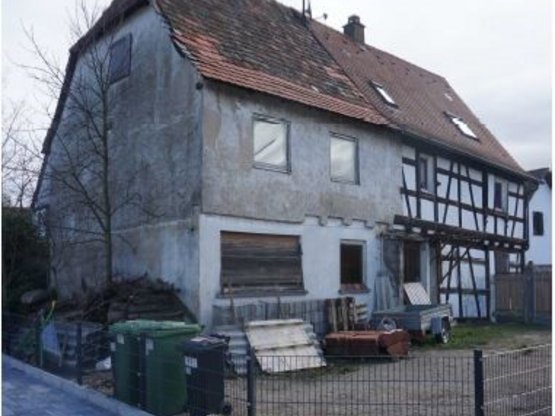 Fachwerkhaus mit Hinterhaus. Hinter dem Gartenzaun stehen Mülltonnen, vor dem Hinterhaus lagern Baumaterialien.