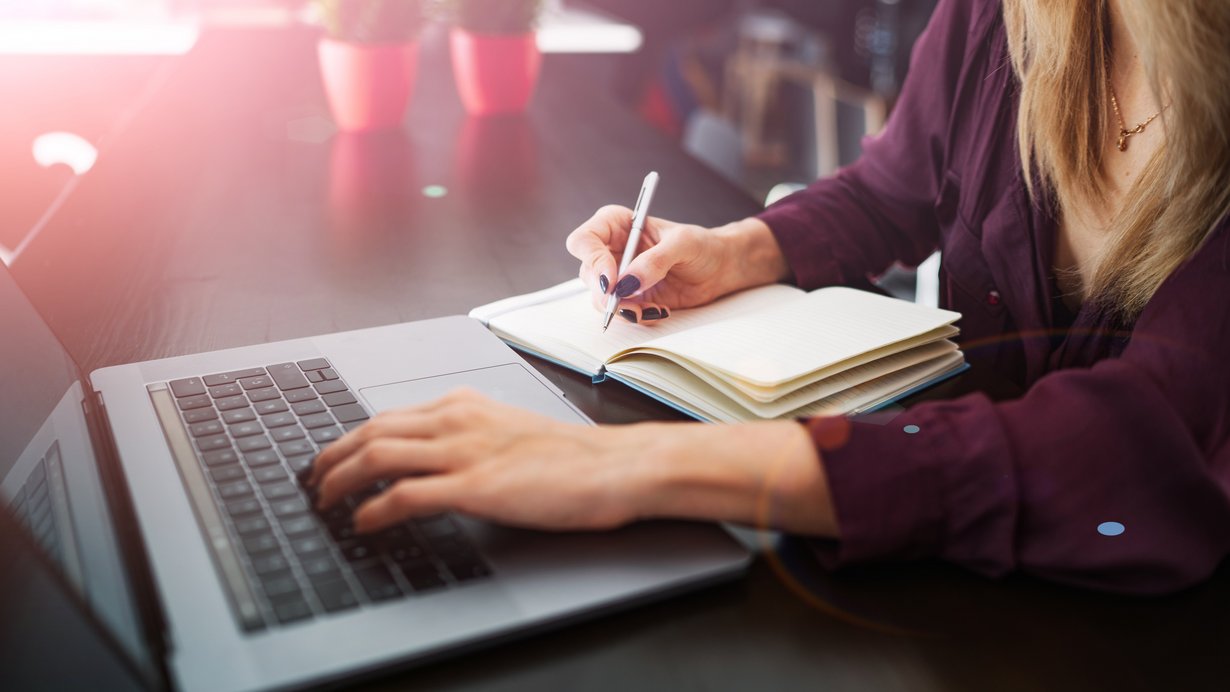 Frauenhand schreibt in ein Notizbuch, während auf dem Laptop im Internet gesurft wird