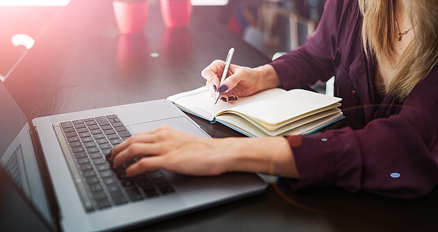 Frauenhand schreibt in ein Notizbuch, während auf dem Laptop im Internet gesurft wird