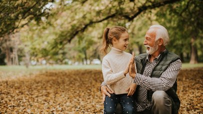 Ein älterer Mann und ein junges Mädchen knien im Freien auf dem Boden und klatschen sich in die Hände. Hinter ihnen ist ein Wald zu sehen.