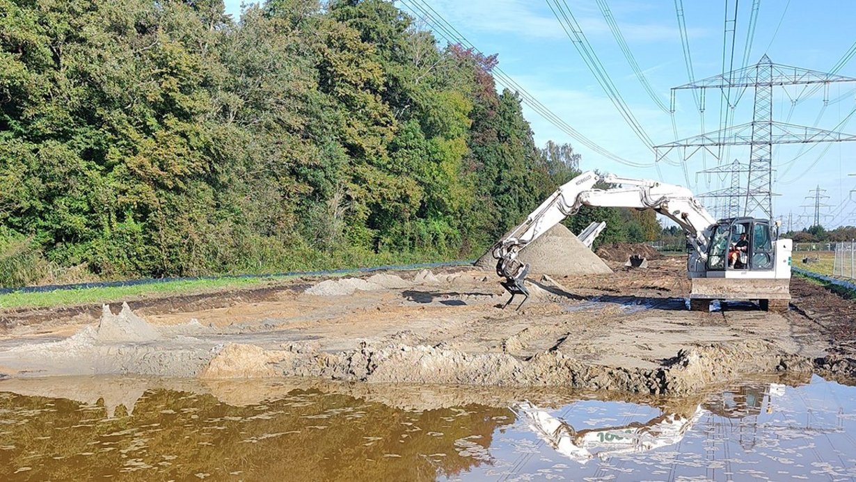 Bagger, der ein großes Loch ausgehoben hat, das inzwischen mit Wasser gefüllt ist. Im Hintergrund der Baustelle befindet sich auf einer Seite Wald, auf der anderen Seite Hochspannungsleitungen. 