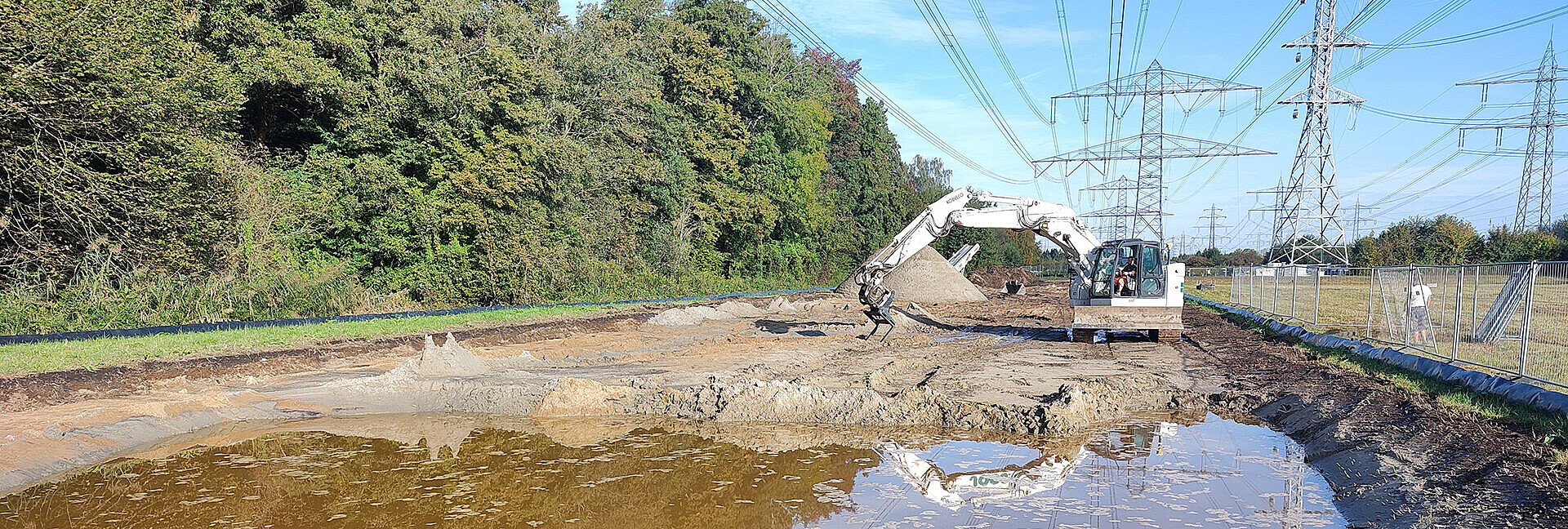 Bagger, der ein großes Loch ausgehoben hat, das inzwischen mit Wasser gefüllt ist. Im Hintergrund der Baustelle befindet sich auf einer Seite Wald, auf der anderen Seite Hochspannungsleitungen. 