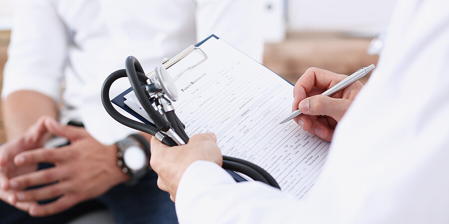 Bild zeigt einen Arzt und Patient, der Arzt hält ein Stetoskop und einen Schreibblock in der linken Hand, schreibt mit der rechten Hand auf den Block