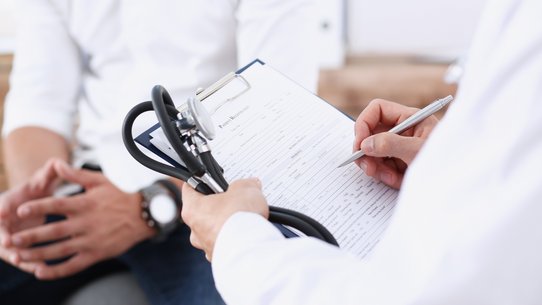 Bild zeigt einen Arzt und Patient, der Arzt hält ein Stetoskop und einen Schreibblock in der linken Hand, schreibt mit der rechten Hand auf den Block