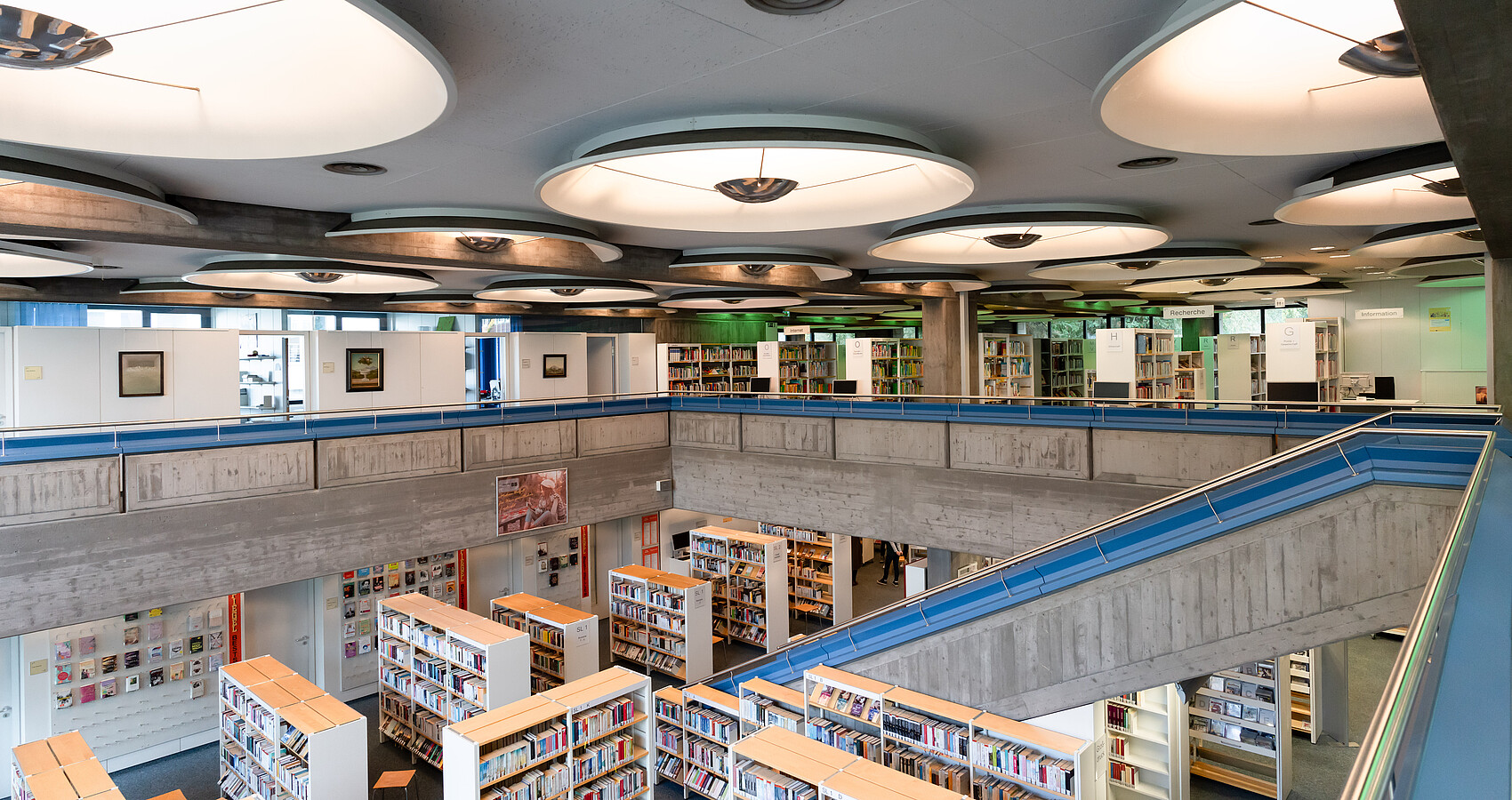 Stadtbibliothek fotografiert vom oberen Stockwerk, rechts eine Treppe, die nach unten führt, überall Bücherregale mit Büchern