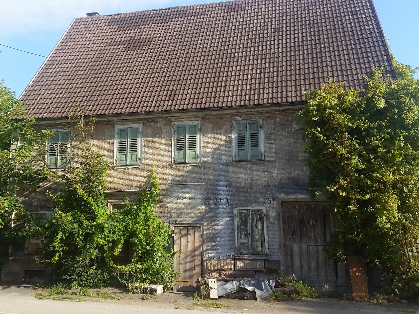 Das Bild zeigt die Frontansicht eines alten, offensichtlich unbewohnten Hauses. Die Fensterläden sind geschlossen, vor dem Haus wuchern große Gebüsche. 