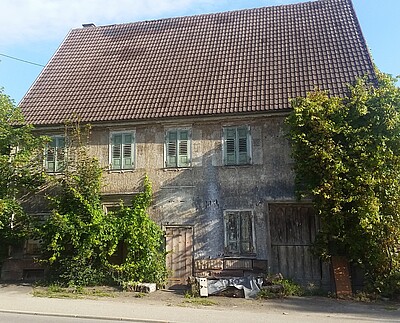 Das Bild zeigt die Frontansicht eines alten, offensichtlich unbewohnten Hauses. Die Fensterläden sind geschlossen, vor dem Haus wuchern große Gebüsche. 