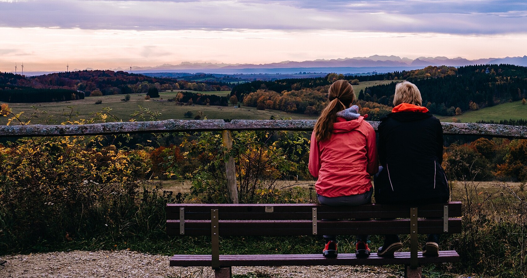 Zwei Frauen sitzen bei Sonnenaufgang auf einem Berg auf einer Bank und schauen ins Tal