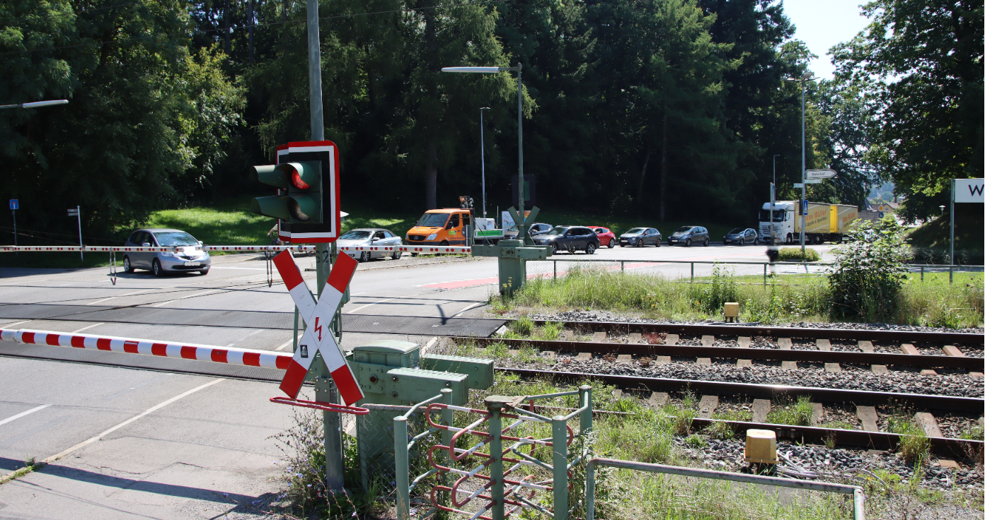 Das Bild zeit einen Bahnübergang mit heruntergelassenen Schranken und eine rote Ampel. Mehrere wartende Fahrzeuge