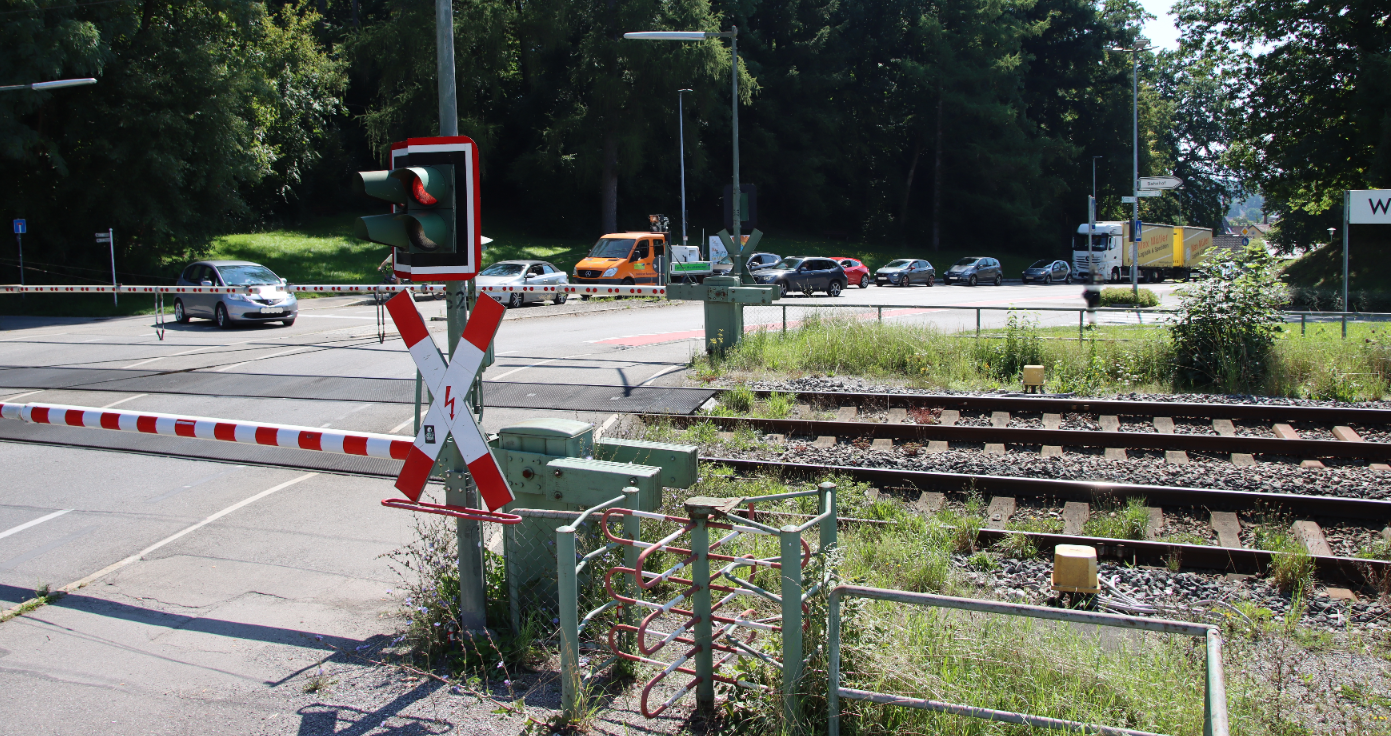 Das Bild zeit einen Bahnübergang mit heruntergelassenen Schranken und eine rote Ampel. Mehrere wartende Fahrzeuge