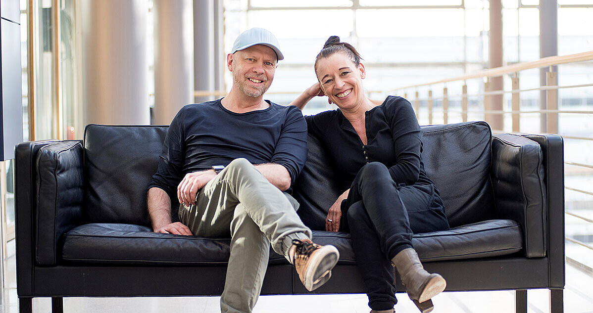Andreas Hüging und Angelika Niestrath sitzen auf einem schwarzen Ledersofa. 