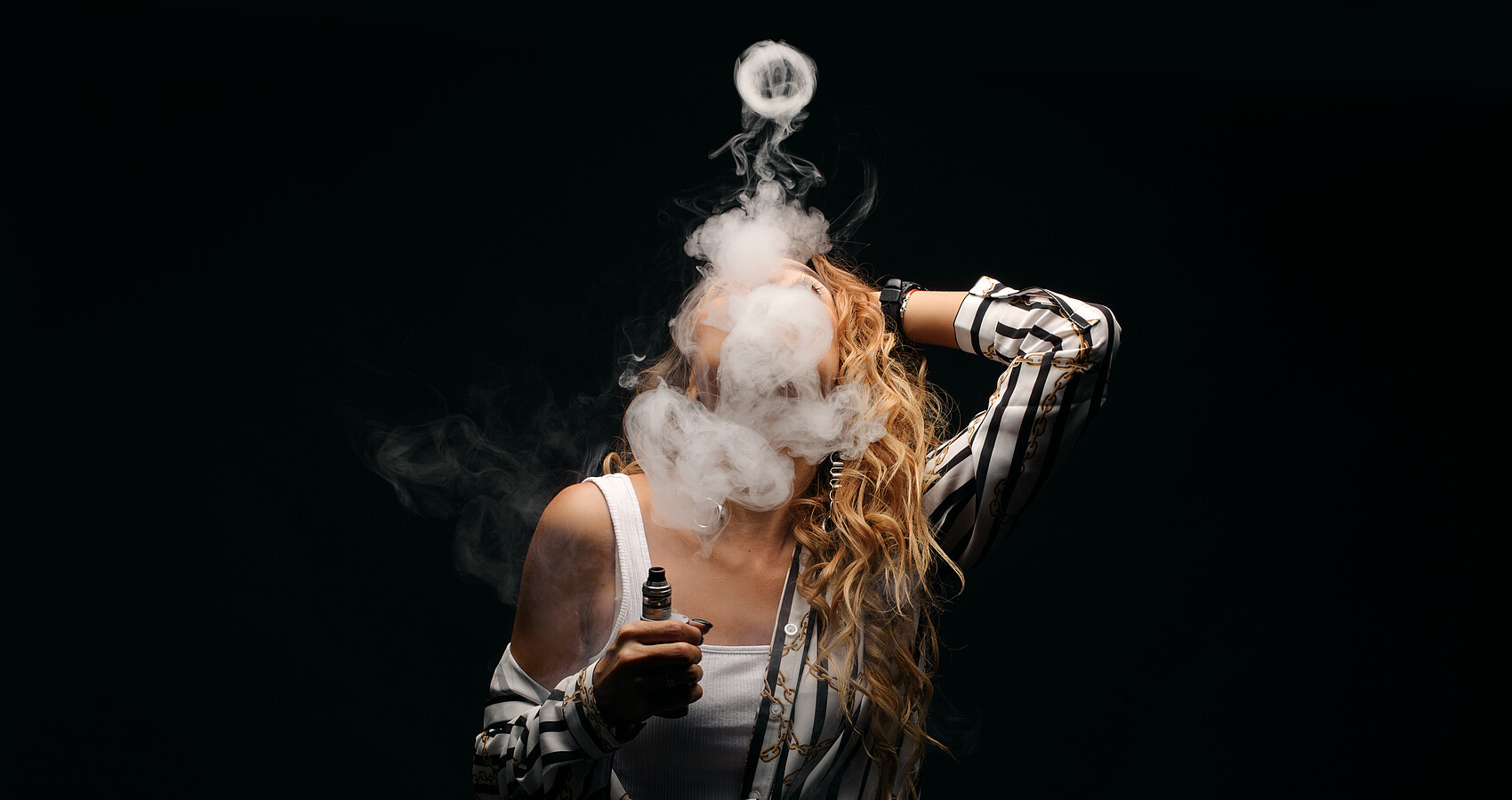 Das Foto zeigt eine rothaarige Frau mit einer E-Zigarette und viel Rauch vor dem Gesicht vor einem dunklen Hintergrund in einer Nahfaufnahme.
