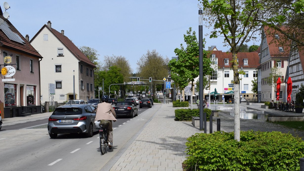 Foto einer Ortsmitte. Neben der Straße, auf der mehrere Autos fahren, befindet sich ein Radstreifen mit einem Radler und ein gestalteter Platz mit Bäumen und Sitzgelegenheiten