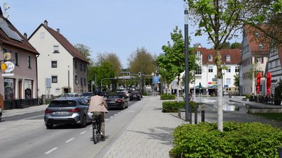 Foto einer Ortsmitte. Neben der Straße, auf der mehrere Autos fahren, befindet sich ein Radstreifen mit einem Radler und ein gestalteter Platz mit Bäumen und Sitzgelegenheiten