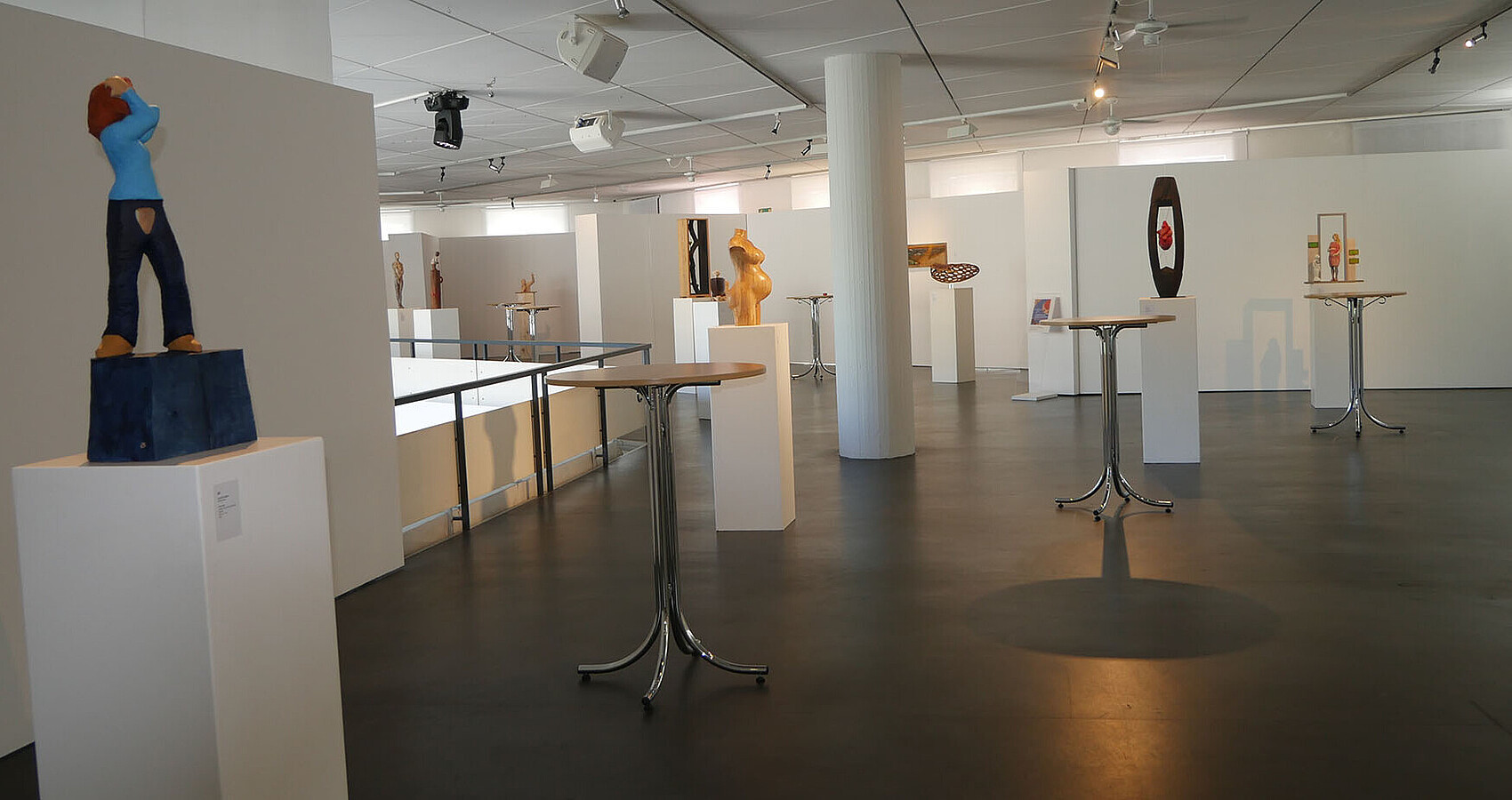 Das Bild zeigt einen Ausstellungsraum, in dem verschiedene Figuren und Skulpturen auf weißen Podesten stehen.