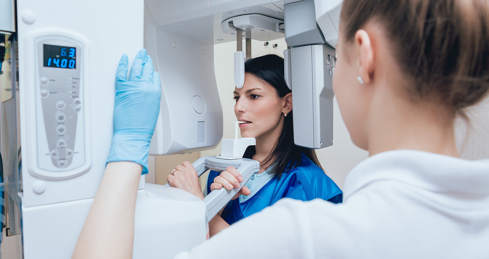 Eine Frau steht in einem Röntgenapparat, bereit für die Aufnahme. Sie trägt einen Strahlenschutz. Eine Arzthelferin macht die letzten Einstellungen am Gerät.Eine 